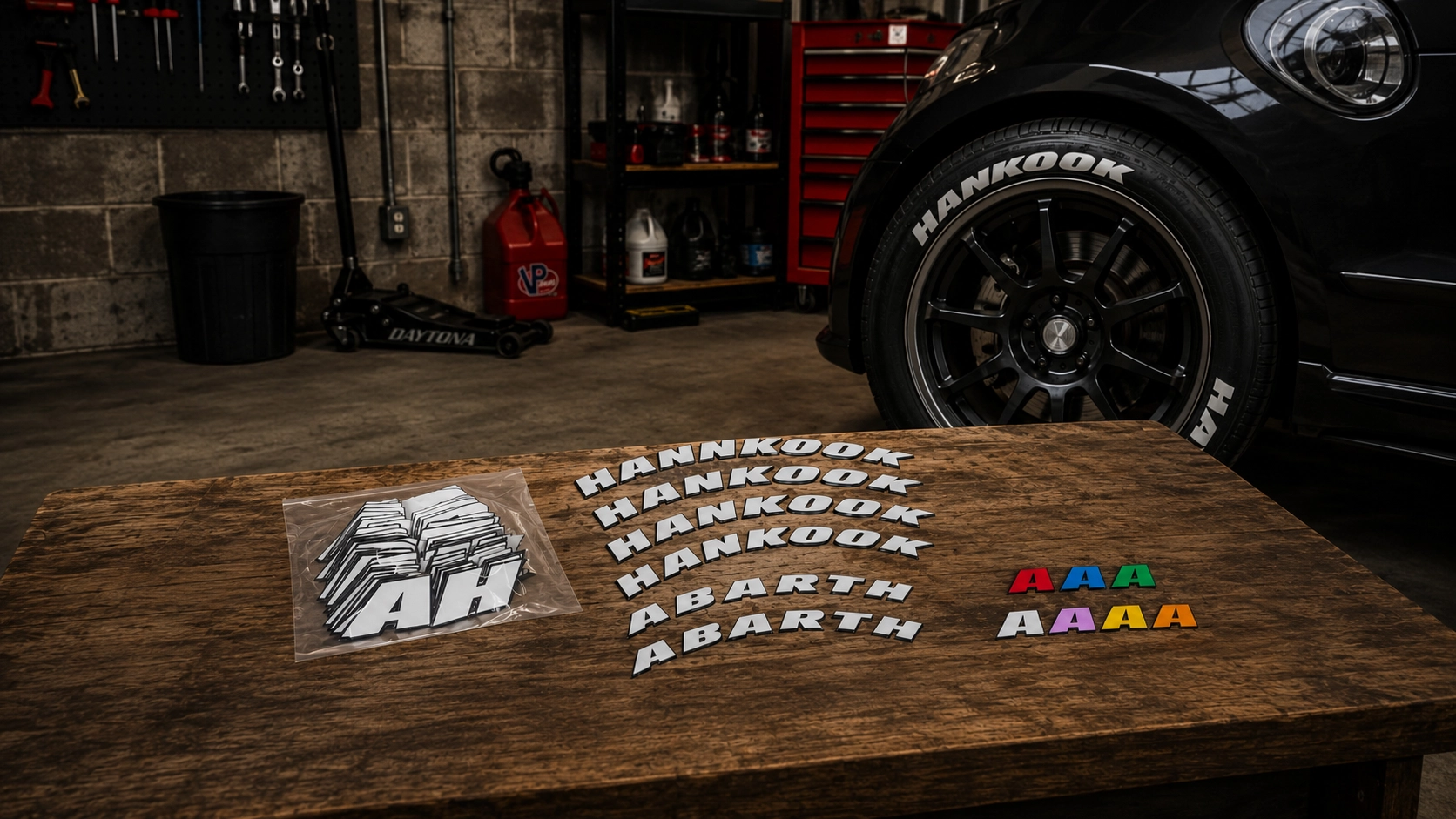 Tire lettering sets on a garage workbench — HANKOOK and ABARTH raised letters beside a car fitted with white sidewall lettering